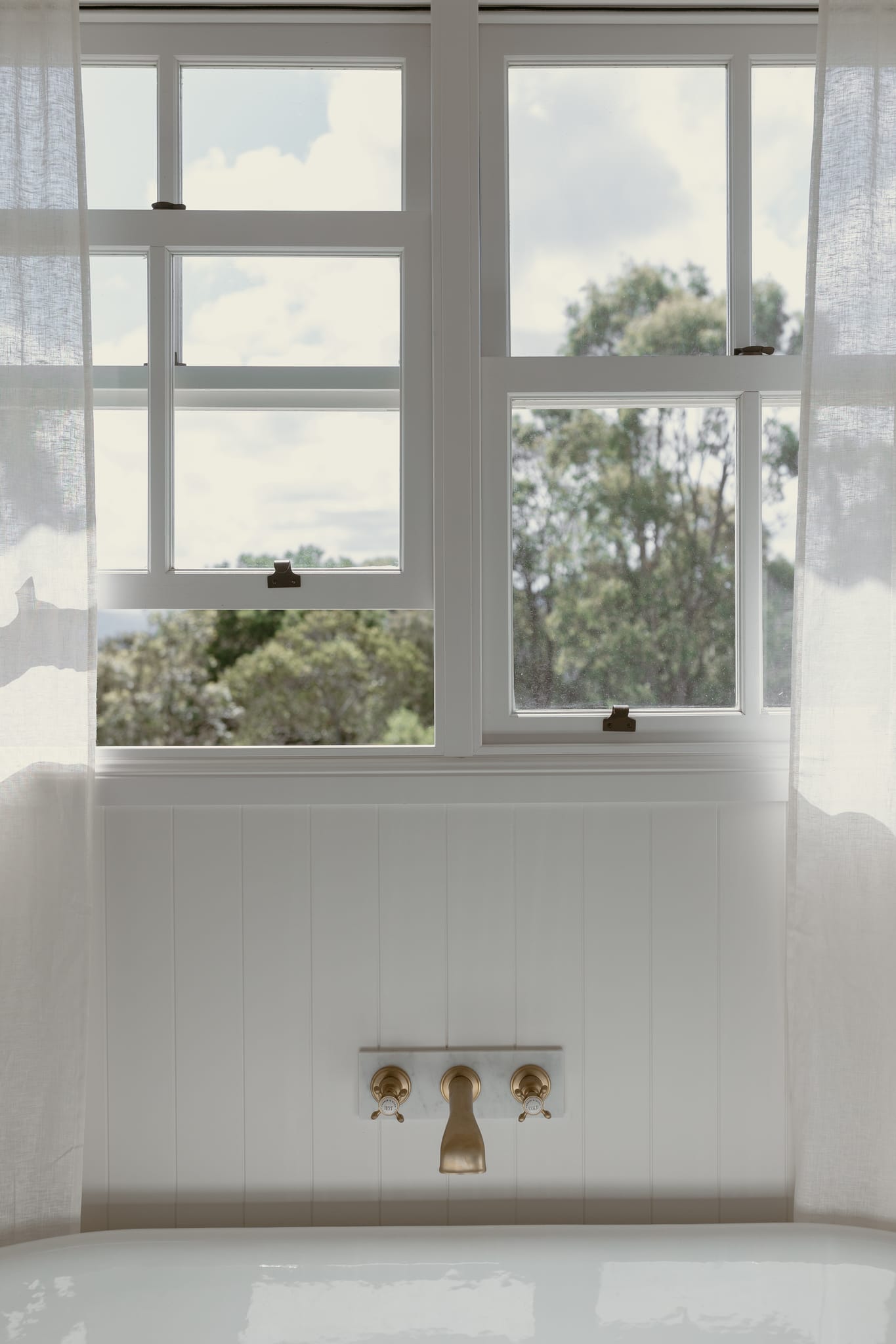 White timber sash windows fitted with sash lifts above a bathtub with brass tapware